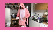 Screenshot: Ein Musiker der NDR ElphCellisten steht im Flamingo-Anzug mit einer Toilettenpapierrolle in einer Küche. © NDR 