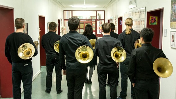 Hornisten des NDR Sinfonieorchesters stehen mit dem Rücken zur Kamera in einem Schulflur.  
