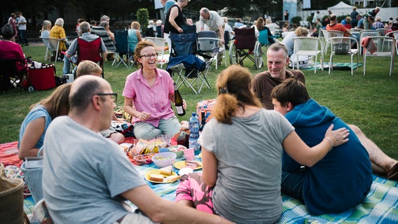 Eine Gruppe von Hannover-Proms-Fans stößt vor Beginn der Veranstaltung mit Wein an, während alle auf der Picknickdecke sitzen. © NDR Foto: Julius Matuschik