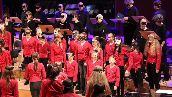 Viele Sänger, alle in Rot oder Schwarz gekleidet, alle mit Augenmaske. © NDR Foto: Thomas Becker