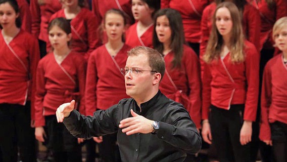 Dirigent wendet jungen Sängerinnen Rücken zu, dirigiert ins Publikum. © NDR Foto: Thomas Becker