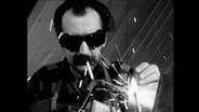 Skulpteur Mário Cravo Junior mit Sonnenbrille und Zigarette beim Schweißen an einer Plastik (1964)  