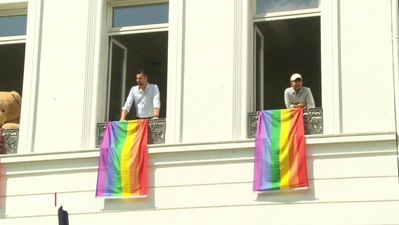 Zwei Männer halten eine Regenbogen-Flagge aus dem Fenster. © Screenshot 