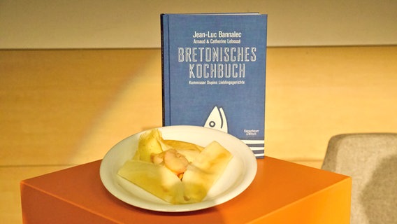 Ein Buch steht neben einem Teller mit einem Gebäck. © NDR 
