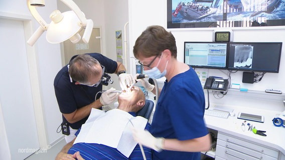 Blick in eine Zahnarztpraxis: einem Patienten wird von einem Arzt in den Mund geschaut, eine Zahnarzthelferin hält einen Schlauch. © Screenshot 