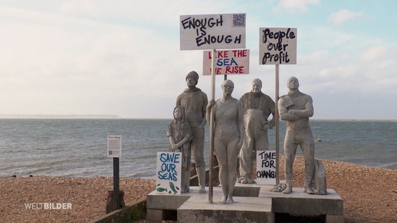 Auf einer Plattform am Strand stehen Skulpturen, die demonstrirenden Menschen nachemfunden sind und halten Protestschilder. © Screenshot 