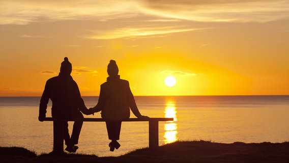 Zwei Menschen in Winterkleidung betrachten Hand in Hand auf einer Bank einen Sonnenuntergang am Wasser. © panthermedia Foto: Ian Lishman