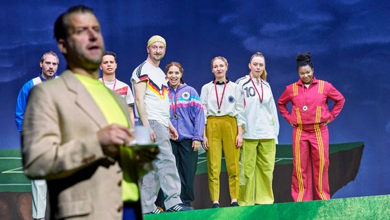 Ein Mann hält eine Kaffeetasse in der Hand, im Hintergrund stehen sieben Männer und Frauen in Sportkleidung. © Staatstheater Hannover Foto: Katrin Ribbe