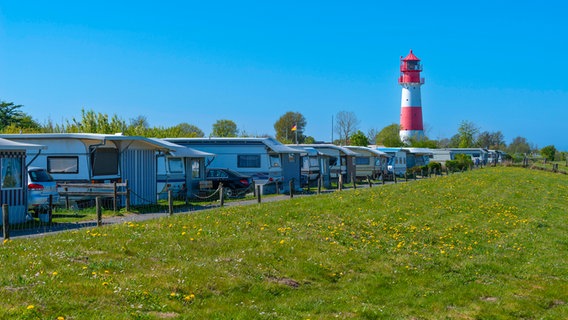 Auf einem Campingplatz stehen mehrere Wohnmobile und Wohnwagen, im Hintergrund steht ein Leuchtturm © picture alliance / imageBROKER Foto: Wolfgang Diederich