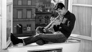 Chet Baker sitzt am offenen Fenster auf Fensterbank und spielt Trompete, Schwarzweiß-Foto © picture-alliance/akg-images / Bianconero 