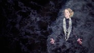 Vor einem schwarzen Hintergrund steht rechts im Bild eine Frau mit schwarzem Kleid und vielen langen Perlenketten um den Hals. © Ernst Deutsch Theater Foto: Oliver Fantitsch
