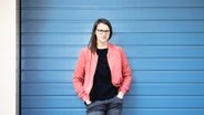 Eine junge Frau mit langem breunem Haar und Brille in einer rosa Jacke vor einer blauen Wand. © Sebauer.Filmer - DuMont Verlag Foto: Sebauer.Filmer