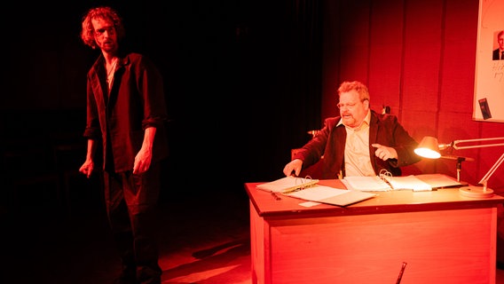 Auf einer Bühne sitzt ein Mann in einer Art Amtsstube hinter einem Schreibtisch. Davor steht ein anderer, jüngerer Man nmit lockigem haar. Die Bühen ist in rotes Licht getaucht. © Theater Das Zimmer Foto: Patrick Bieber