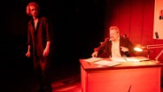 Auf einer Bühne sitzt ein Mann in einer Art Amtsstube hinter einem Schreibtisch. Davor steht ein anderer, jüngerer Man nmit lockigem haar. Die Bühen ist in rotes Licht getaucht. © Theater Das Zimmer Foto: Patrick Bieber
