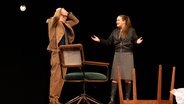 Auf einer Bühne stehen ein Mann im braunen Cordanzug und eine Frau mit Zopf und Lederjacke. Der Mann hebt die Hände über den Kopf und die Frau breitet die Arme aus und redet offenbar auf den Mann ein. © St. Pauli Theater Foto: Kerstin Schomburg