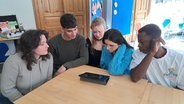 Fünf junge Menschen sitzen um einen Tisch herum. Sie schauen auf ein ipad. Es liegt auf dem Tisch in der Mitte. © NDR Foto: Petra Volquardsen
