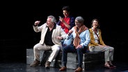 Auf schwarzen Kisten auf einer Bühne sitzen mehrere Schauspieler und Schauspielerinnen. © G2 Baraniak Foto: G2 Baraniak