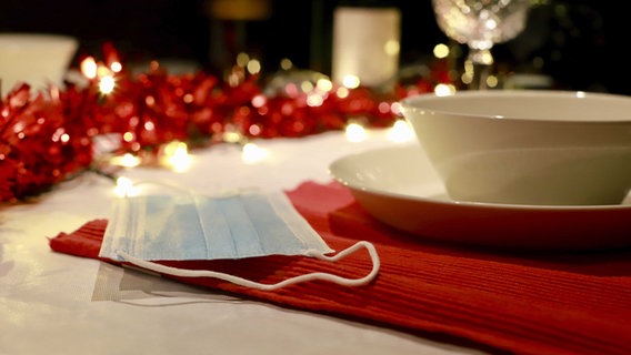 Eine Mund-Nasen-Bedeckung liegt auf einem Tisch neben einem Teller. Im Hintergrund ist Weihnachtsdekoration zu sehen. © picture alliance/Richard Brocken/ANP/dpa Foto: Richard Brocken