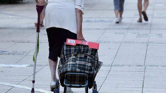 Eine alte Frau mit einer Krücke und Einkaufstrolley geht durch eine Fußgängerstraße. © picture alliance/dpa/dpa-Zentralbild/Jens Kalaene 