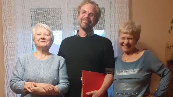 Ein Mann mit einem Buch in der Hand steht zwischen zwei älteren Damen. © privat 