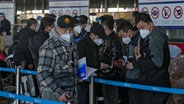 Reisende mit Mund-Nasen-Schutz überprüfen ihre Pässe, während sie am Schalter für internationale Flüge am Flughafen Peking anstehen. © AP/dpa Foto: Andy Wong