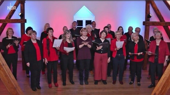 Rot gekleidete Damen und Herren mittleren Alters halten Notenhefte und singen. © NDR Kultur 