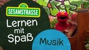 Elmo steht vor einer Wand mit Graffiti und hinter einem braunen Klavier. Neben ihm steht ein Schild auf dem steht: "Sesamstraße: Lernen mit Spaß - Musik". © NDR 