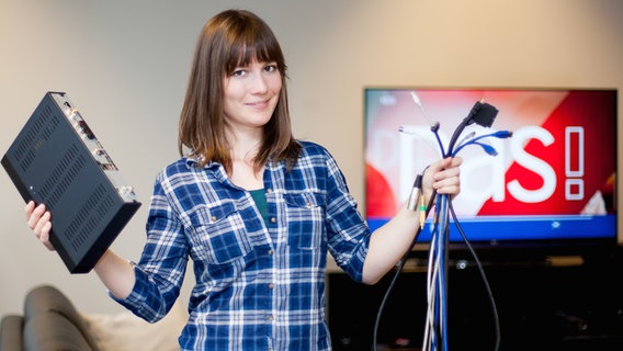 Eine Frau steht mit einem Bündel Kabel und einem Receiver in den Händen vor einem Fernseher © NDR Foto: Patrick Joosten