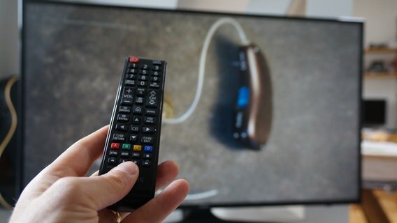 Eine Fernbedienung wird vor einen Fernseher gehalten, auf dem ein Hörgerät zu sehen ist. © NDR Foto: Moritz Kirscht
