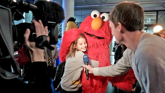 Präsentation der Sesamstraßen-Sondermarke: Ein Mädchen umarmt Elmo ganz fest, während ein Kameramann es filmt und es von einem NDR Reporter interviewt wird. © NDR/Jann Wilken Foto: Jann Wilken