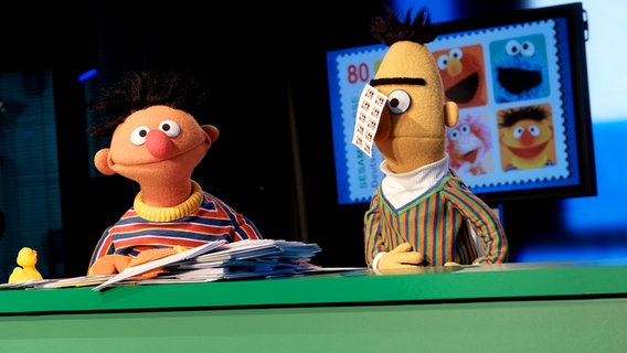Bei der Präsentation der Sesamstraßen-Briefmarke: Ernie und Bert hinter einem grünen Tisch. An Berts Nase klebt ein Bogen mit den neuen Briefmarken. © NDR/Jann Wilken Foto: Jann Wilken