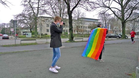 Zwei Mediengestalter vor dem Landesfunkhaus Hannover: eine Person fotografiert die andere, die eine Regenbogenflagge wie einen Umhang trägt. © NDR 