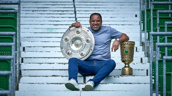 Der ehemalige Fußballspieler Ailton sitzt mit einer Meisterschale und dem DFB-Pokal auf einer Treppe. © NDR/Radio Bremen/Andreas Gumz Foto: Andreas Gumz