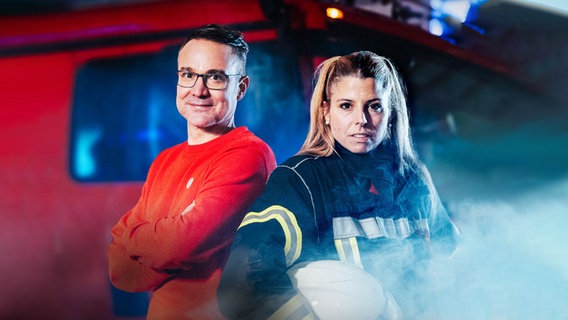 Ein Mann in rotem Pullover und eine Frau in Feuerwehruniform mit Helm stehen vor einem roten Feuerwehrauto mit Blaulicht und Rauch im Hintergrund. © NDR Foto: Andrea Seifert