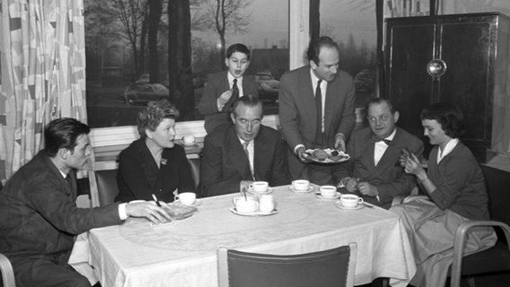 Familie Schölermann sitzt mit zwei weiteren Herren am Kaffeetisch. Einer der Herren bietet Herrn Schölermann eine Kuchenplatte an © NDR 