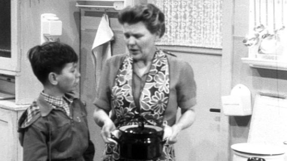 Sohn Jockeli und Mutter Schölermann stehen in der Küche, sie hat einen Kochtopf in der Hand und schaut ihren Sohn an © NDR 