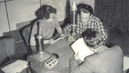 Zwei Männer und eine Frau sitzen in einem Radiostudo an einem Tisch vor Mikrofonen.  