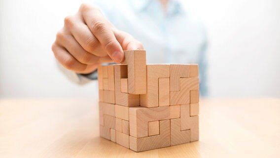 Aus mehreren unterschiedlich geformten Holzstücken ist ein etwa Würfel zusammengesetzt worden. Eine Hand setzt das letzte Stück ein. © Jakub Krechowicz | Getty Images/iStockphoto 