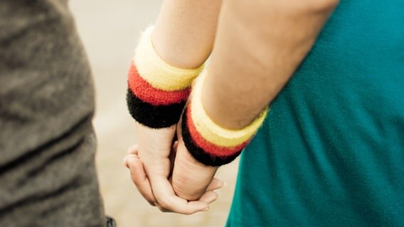 Zwei Menschen mit Frottearmbändern in Schwarz-Rot-Gold halten sich die Hände. © photocase Foto: andrey-fo