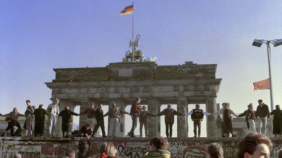 Eine historische Aufnahme von Menschen, die auf der Berliner Mauer stehen, sich die Hände halten und die Öffnung der Grenze feieren. © picture alliance Foto: AP Photo Thomas Kienzle