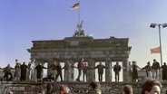 Eine historische Aufnahme von Menschen, die auf der Berliner Mauer stehen, sich die Hände halten und die Öffnung der Grenze feieren. © picture alliance Foto: AP Photo Thomas Kienzle