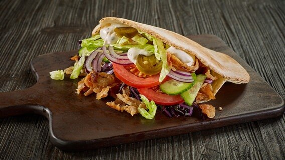 Ein fertig zubereiteter Döner Kebab liegt auf einem dunklen Hozbrett: Ein aufgeschnittenes Fladenbrot mit Zwiebeln, Tomaten, Salat, Fleisch und Sauce. © Colourbox 
