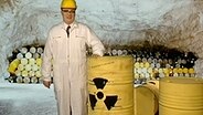 Ein Mann im weißen Overall steht neben einem gelben Faß mit dem Atomzeichen  