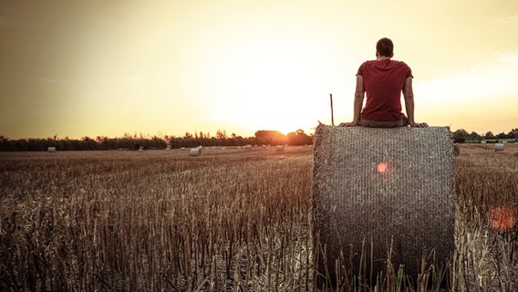 Ein Mann sitzt auf einem Strohballen auf einem Acker und betrachtet den Sonnenuntergang. © David-W- / photocase.de 