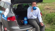 Andreas Scheuer bläst im Kofferraum sitzend einen Luftballon auf.  