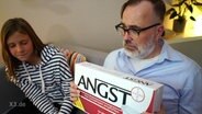 Ein Mann und ein Mädchen auf einem Sofa. Er hält eine überdimensionierte Medikamentenschachtel in der Hand. Aufschrift:  ANGST  