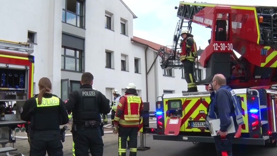 Polizisten und Feuerwehrleute stehen vor einem Feuerwehrauto und blicken auf ein weißes Haus. © Nord-West-Media TV & Nachrichten GmbH 