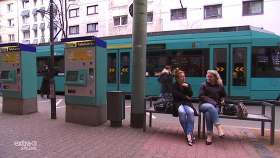 Zwei Frauen sitzen auf einer Bank mit dem Rücken zur Straßenbahn.  