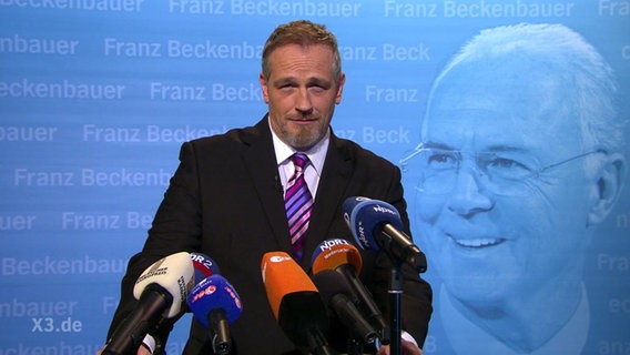 ein Mann vor Mikrophonen, im Hintergrund ein Bild von Beckenbauer  