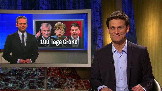 Der Moderator Christian Ehring. Im Hintergrund ist ein Bildschirm mit der Aufschrift "100 Tage GroKo" zu sehen.  